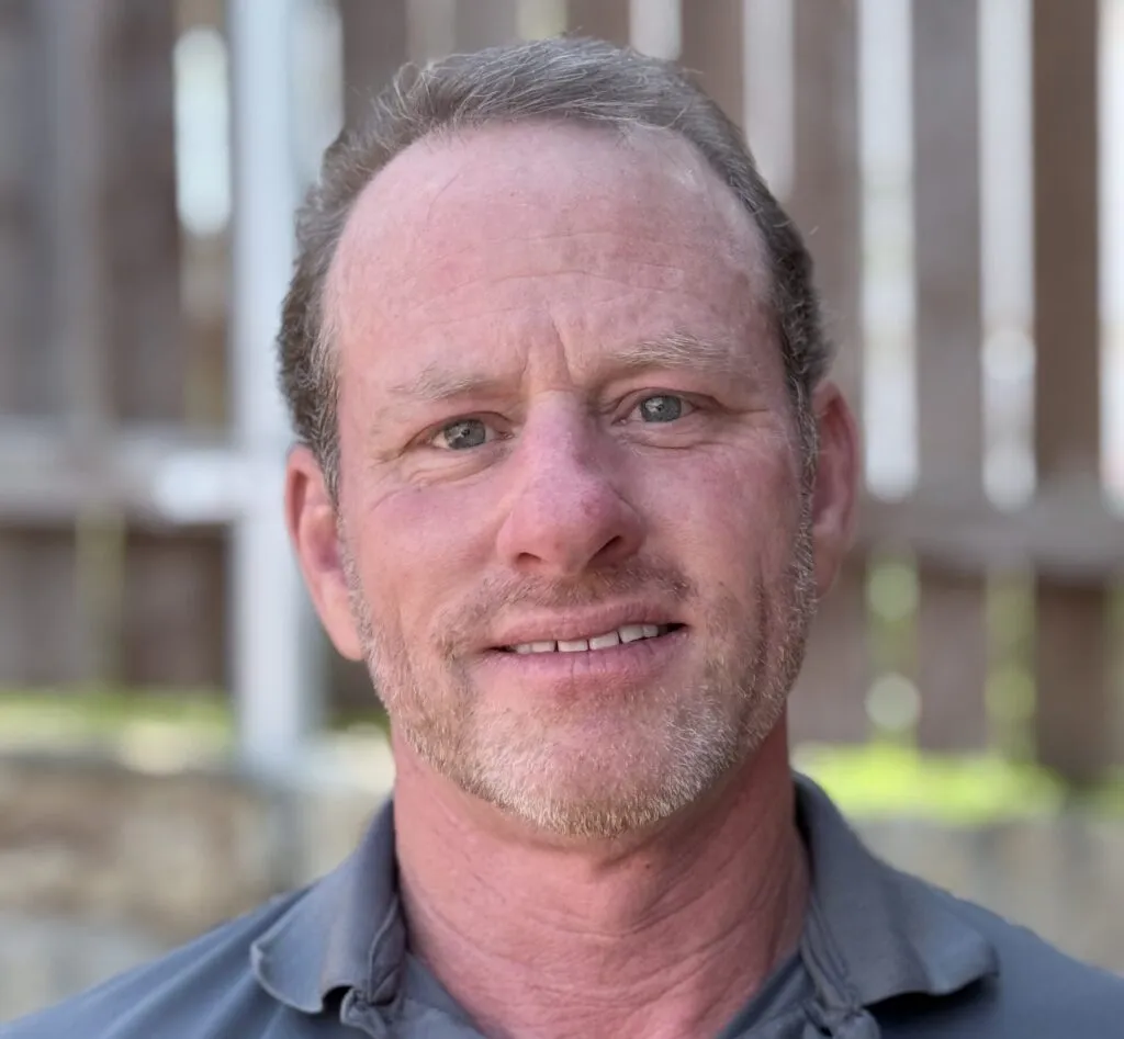Professional HVAC technician smiling, representing Kelly's Heating and Air, with a blurred wooden fence in the background, emphasizing expert care and customer service in HVAC solutions.