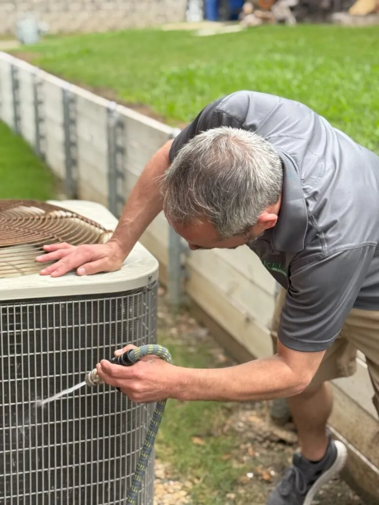 Man cleaning AC unit with hose in Lewisville, TX. Air conditioning maintenance ensures efficient cooling and extends the system's lifespan.