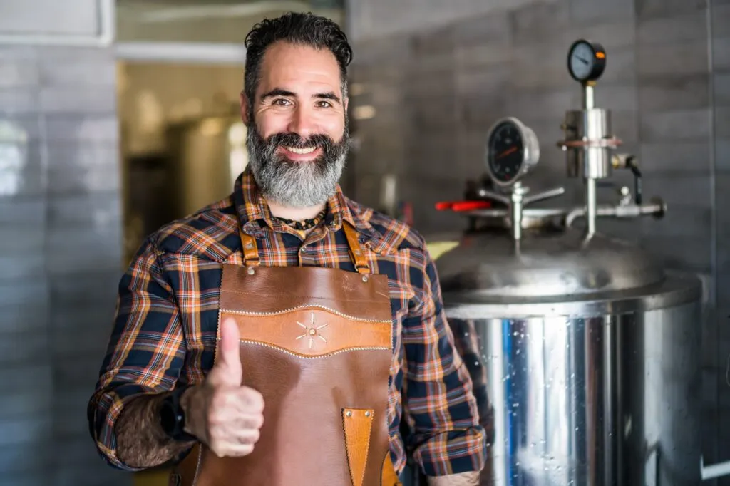 Brewmaster giving a thumbs up in front of brewing equipment. Man with beard wearing an apron. Craft beer brewing process in Lewisville, TX.